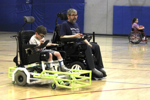 A man and child in power soccer wheelchairs on an indoor court.