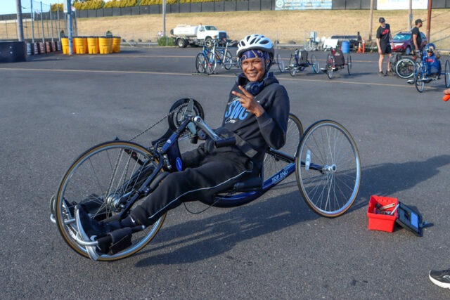 Women smiling and giving peace sign in a hand cycle with tool box on the ground nearby.