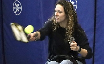 Action shot of a woman seated in a wheelchair using a paddle to play pickleball.
