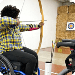 Woman seated in a wheelchair using a recurve bow aimed at a target on a haystack wall.