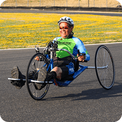 Individual using a seated handcycle wearing sunglasses and a cycling helmet on a racetrack.
